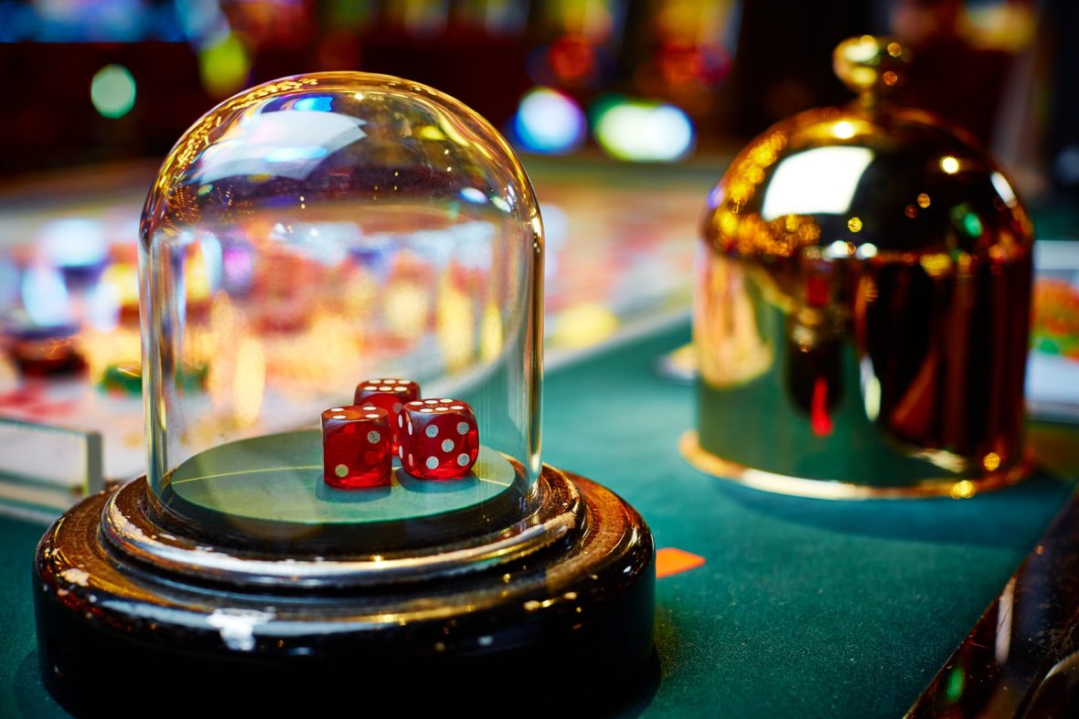 Sic Bo casino game setting showcasing a close-up of a glass dome containing red dice with white dots, resting on a green surface, with blurred colorful lights and a golden bell in the background.